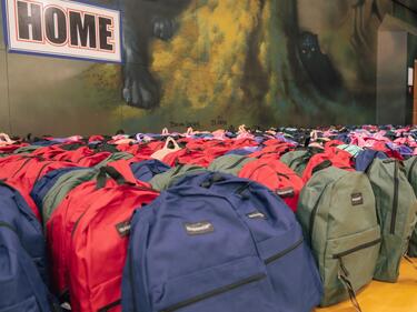 A large number of filled backpacks lined up in rows up against a gymnasium wall.