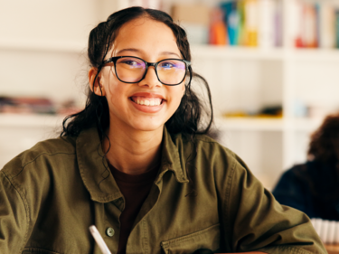 Student wearing glasses sits at a desk and writes on a worksheet in a bright classroom. Bookshelves are visible in the background, and a Fraser Health logo appears in the bottom-right corner of the image.
